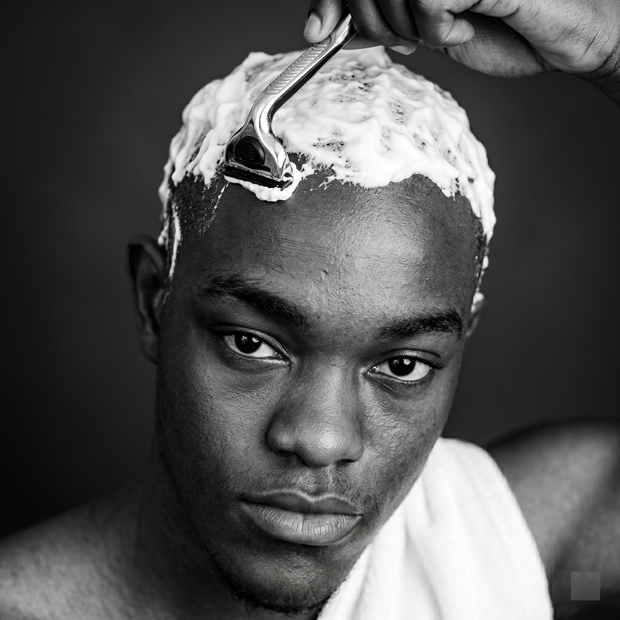 A young man with a towel around his neck is shaving his head while looking directly at the camera. He has shaving cream applied to his scalp.